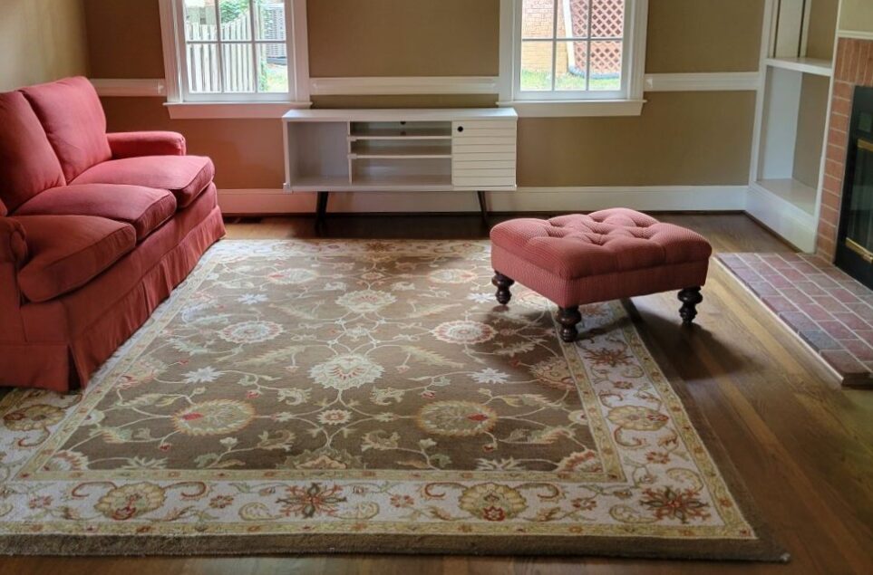 The back LR rug that the previous owners of the house sold to us, with a generic traditional motif and uninspiring colours of dull browns, tans and a bit of maroon to match a garish maroon couch (seen to left). A matching brick red tufted footstool stands to right (to accompany an oversized stuffed arm chair of same colour that's been relegated to the basement, also sold by previous owners). Straight ahead is south-facing wall with two windows and our white midcentury-modern console table in between (having moved in two days prior, no TV screen or other furnishings are yet unpacked). To the right and mostly out of sight is fireplace along wall shared with the front LR, and built-in bookcase in corner. The walls are painted a split-pea soup murky brown to match the rug.