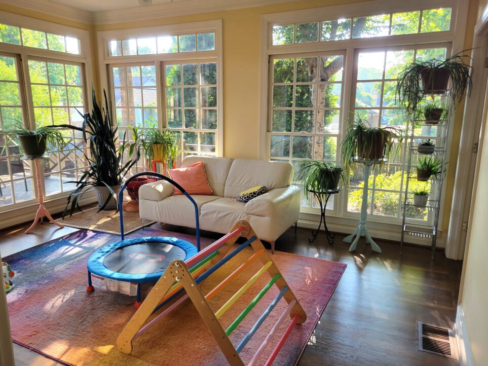 The sunroom viewed five days after moving (the only truly peaceful and put-together room in the house at the time): straight ahead is a wall of floor-to-ceiling windows facing east, letting in plenty of natural light for eleven plants and counting (more are out of view) positioned on various plantstands, small tables, and shelves along that wall as well as the north-facing wall to the left with its continuing floor-to-ceiling windows. In the center of the floor is a colourful ombre area rug with a small children's trampoline and colourful Pikler triangle in the middle.