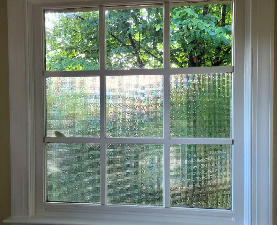 One of the two square-shaped primary bathroom windows, which (along with all windows in this house) is divided into smaller sections by removable white-painted wooden 'window pane dividers'. The lower six sections are covered in a stick-on iridescent/clear film to give privacy whilst still letting in natural light. In the above three sections, the glass is bare so treetops are still seen unobscured (but high enough for privacy from people in yards/houses behind)