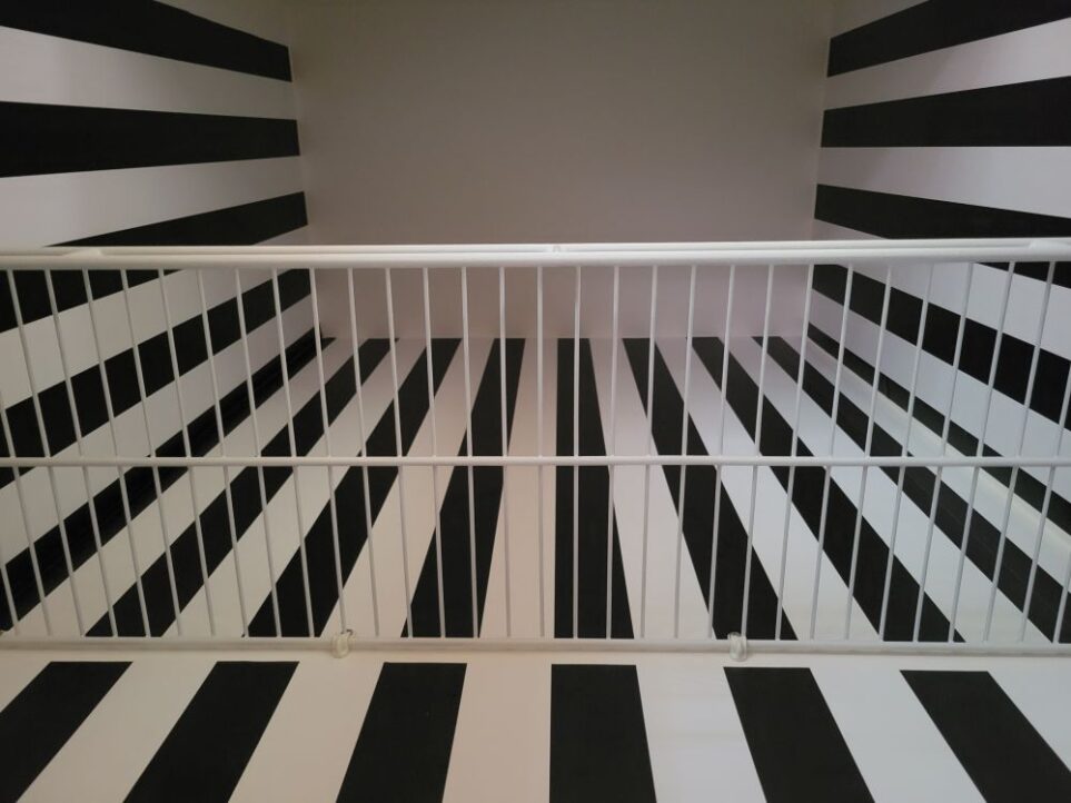 Small white ceiling of empty coat closet viewed from below white rubber-coated wire shelf: the three walls are covered in vertical black-and-white striped peel-and-stick wallpaper