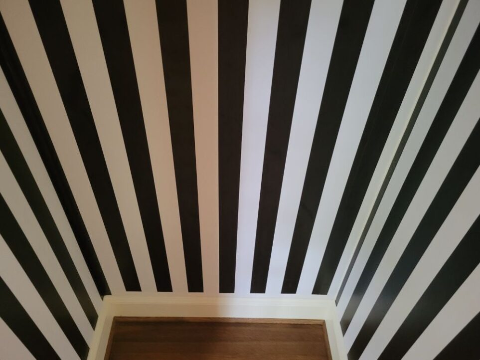 Small wood floor of empty coat closet viewed from inside, and showing three walls covered in vertical black-and-white striped peel-and-stick wallpaper