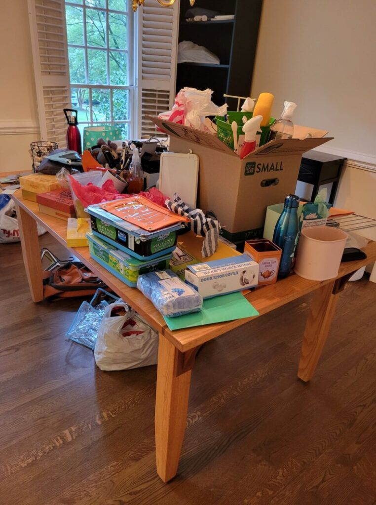 The table in the dining room is covered in stuff five weeks after moving house: an open moving box filled with cleaning supplies sticking out, Swiffer wipes, metal water bottles, bins, childproofing materials, decorations, folders, small boxes, etc.
