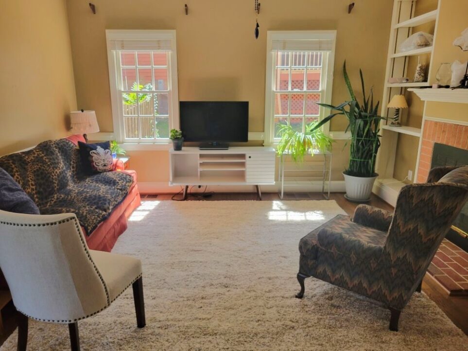 Back LR, still sparsely furnished two months after move but looking much improved with several houseplants near windows, end tables and lamps, and new (used) chairs that are more attractive. A leopard-print throw blanket covers the maroon couch lining left wall. Instead of offensive wool rug which has been removed (offensive both in look and smell) is a new light-coloured off-white/beige high-pile area rug covering most of the LR floor, that is both more attractive and much more comfortable (and has no odor, most importantly!)