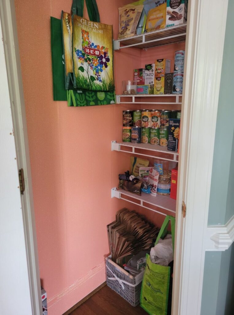 The open pantry stocked with organised dry goods and a few reusable grocery bags hanging from hook on wall. The walls are covered in a sparkling peach peel-and-stick wallpaper. On the floor is a basket with paper bags