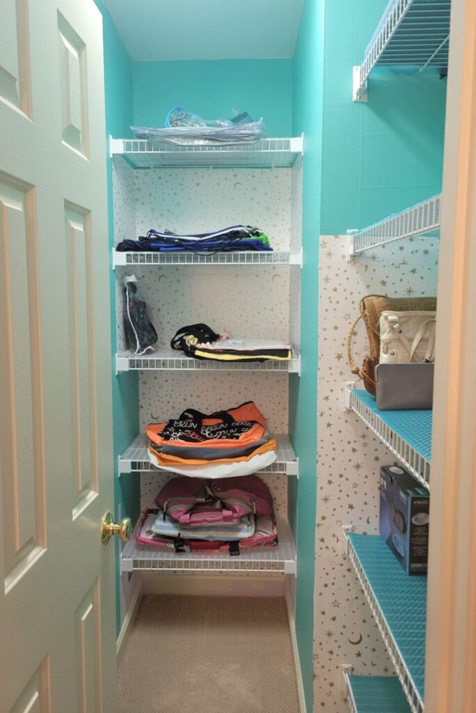 A view of the walk-in closet in the hallway outside the office door: white rubber-coated wire shelves with transparent blue shelf-liner span part of the wall on the right and the narrow section of back wall straight ahead. On the shelves are organized piles of various types of bags. The walls are covered in two types of peel-and-stick wallpaper, applied in intermittent sections: a bright blue colour and a white with small gold stars and moons
