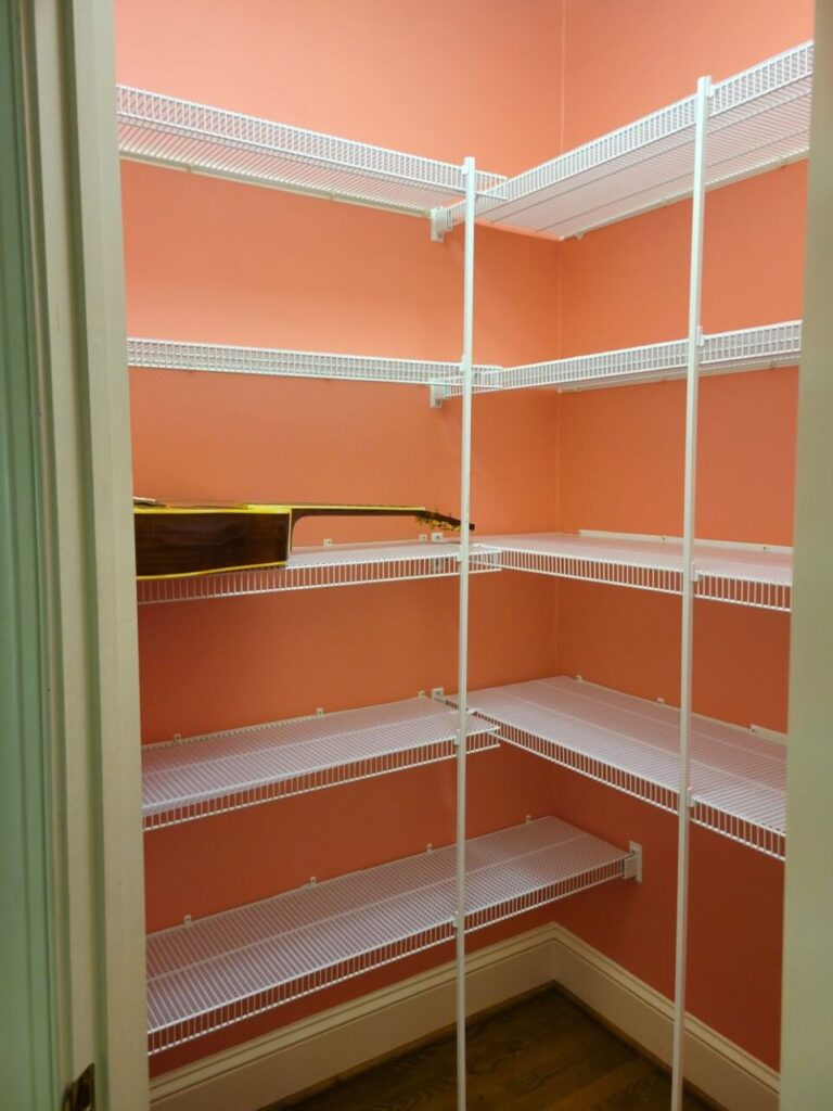 The closet in the study provides ample storage, a must-have to keep creative spaces from getting terribly cluttered. The several white rubber-coated wire shelves, having been recently and thoroughly scoured and cleaned, are now covered in clear shelf liner to prevent future dust and provide a more solid surface on which to set things. The walls are covered in a salmon pink peel-and-stick wallpaper, both to cover many marks and stains and also to create a fun and pretty storage space. A sole guitar rests on one of the shelves, before the shelves were later neatly filled with creative supplies and bins of holiday decorations