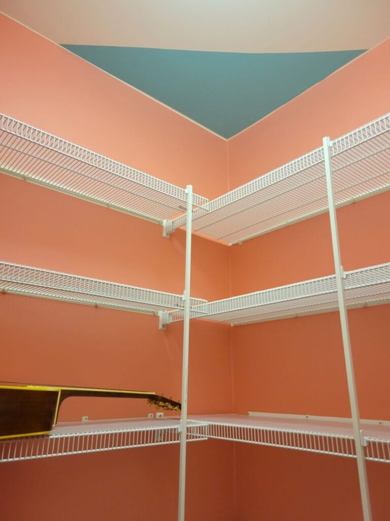 The spacious study closet provides ample storage, a commodity that's very helpful in keeping creative spaces free from clutter. The recently cleaned white rubber-coated wire shelves are newly covered in clear shelf liner. The walls are freshly covered in salmon pink peel-and-stick wallpaper to cover marks and stains and also to create a beautiful storage space. A triangle swath of teal peel-and-stick wallpaper covers a portion of the ceiling, to cover a few more stains there and to complement the pink walls. A guitar is seen resting on one of the shelves before they were later filled with more things.