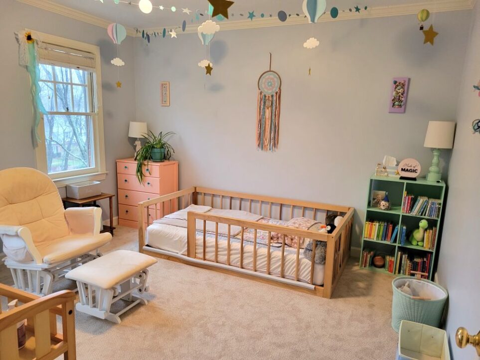 View of baby's room from standing just inside open door. Along opposite wall straight ahead is Montessori floor bed furnished with soft bedding and plush animals. Above it on wall hangs soft colourful dreamcatcher. In right corner next to bed is sea green bookshelf cube cubby with books, plush animals and salt lamp sphere. Atop it is sea green table lamp with white shade, nautical tissue box cover with tissues, and decoration. Above it hangs lavender-framed seashell art. Along wall to right are a small cloth bins on the floor. From ceiling hang paper garlands of sparkly blue stars, as well as blue and lavender paper hot air balloons with clouds and stars. To left of bed and in left corner by window is a peach painted dresser with blue glass drawer knobs, with a white ceramic seahorse lamp and airplane plant on top. Above it hangs a small peach-framed seashell collage. An artificial sunflower with dangling ribbon hangs from corner of window. To left of window is peach glider rocker and stool. Below window is small wood end table with cloth bin on top.