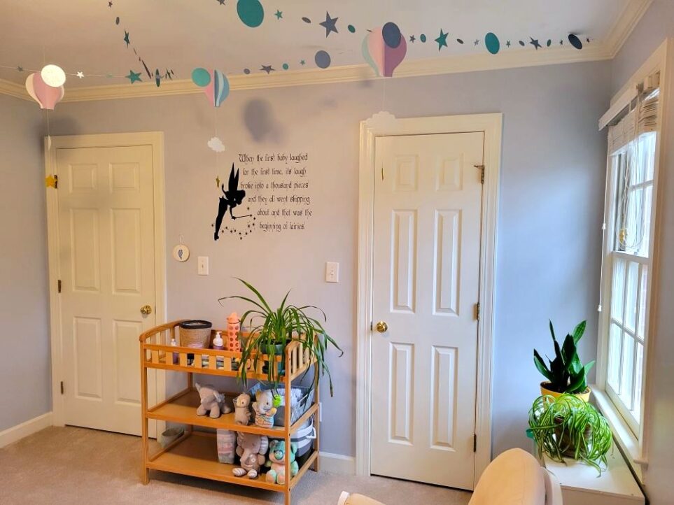 View of baby's room facing wall with closet door and door leading into hallway. In between the two doors is changing table (used as a table) with airplane plant on top along with pull-ups and other toiletries. On shelves below are plush animals. In right corner below window is white cubby table with small snake plant and airplane plant on top. From ceiling hang paper garlands of sparkly blue stars, as well as blue and lavender paper hot air balloons with clouds and stars.