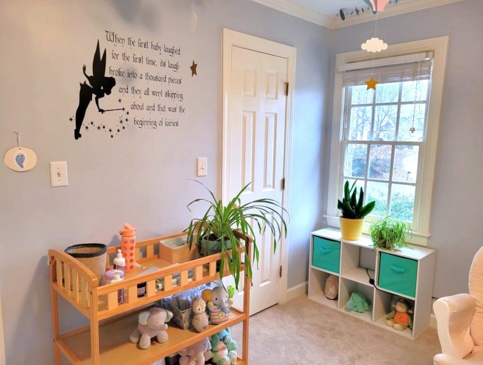 View of baby's room facing corner with closet door and window. Along the wall left of the closet door is changing table (used as a table) with airplane plant on top along with pull-ups and other toiletries. Peter Pan fairies quote decal with Tinkerbell is displayed along wall above it. On lower shelves are plush animals. To the right of the closet door along the adjacent wall, in the corner below the window, is a white cubby table with small snake plant and airplane plant on top. From ceiling hang paper garlands of sparkly blue stars, as well as blue and lavender paper hot air balloons with clouds and stars.
