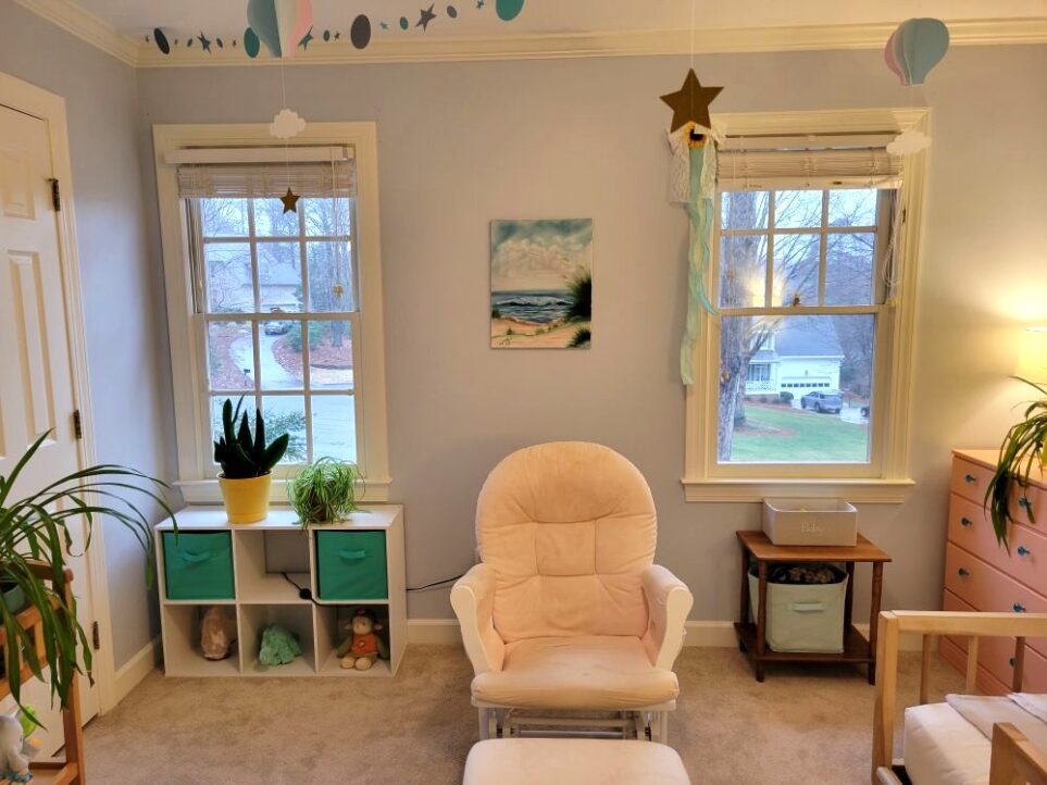 View of baby's room facing wall with two windows and light pink glider rocker in between. Above it hangs an ocean painting on the wall. In the right corner partially out of sight is a peach painted dresser with blue glass drawer knobs, with a small lamp and airplane plant on top partially out of sight. Below right window is a small wood end table with a couple of cloth bins on it. Under window on left is a white cube cubby table turned horizontally with a couple of turquoise cloth bins inside as well as salt lamp and plush animals, and on top is a small snake plant and a small airplane plant. Mostly out of sight on left is closet door and changing table (used as a table) with airplane plant on top. From ceiling hang paper decorations in blue, lavender and gold: stars, clouds, and hot air balloons