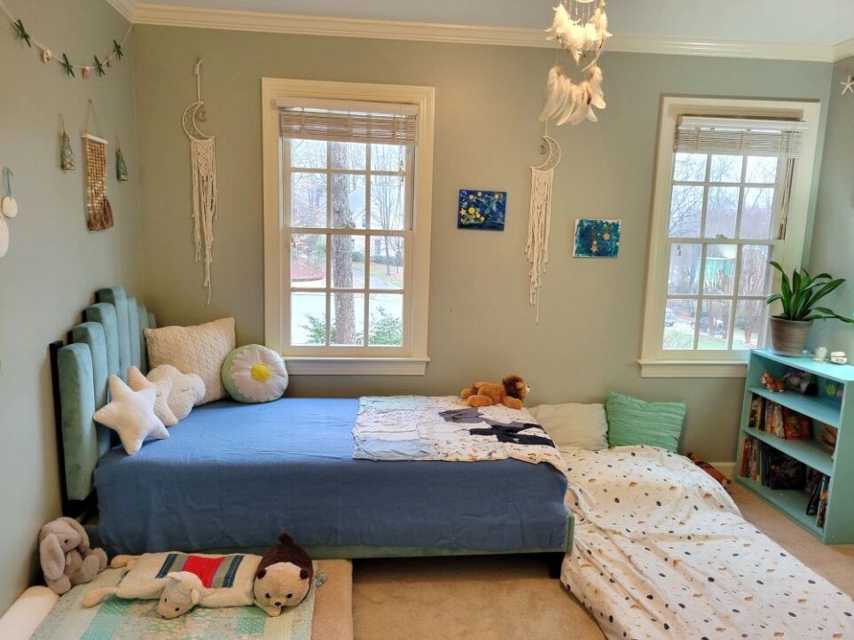 View of the children's bedroom (technically the kindergartener's room but where both boys now sleep at night) facing the wall with two windows. Along the wall in the left corner is a twin bed and blue tufted headboard, covered in star-shaped throw pillows among other pillows and a blue blanket. Lining the left wall and tucked partially under the twin bed is a futon mattress where the toddler sleeps, furnished with plush animals, several little pillows, and matching blanket. Several nautical decorations (mostly out of view) line the wall on the the left, including a seashell/starfish garland above the bed. Above bed on the window wall is a macrame crescent moon dreamcatcher, with a second one hung above the foot of the bed in between the two windows and two blue artworks made by kindergartener. From ceiling light fixture in center of room hangs white feather dreamcatcher. A second twin futon mattress lies on the floor at the foot of the twin bed, with its head against the wall under the left window (where kindergartener sleeps). A small blue-painted bookcase is in the right corner by the right window with a snake plant on top and filled with books.