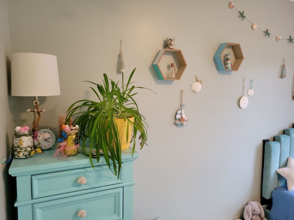 Close-up photo of children's bedroom (technically the kindergartener's room but where both boys now sleep at night) facing the wall with the blue-painted dresser, partially in sight. Atop it is an airplane plant, silver anchor-shaped lamp with white shade, clock, salt lamp, and fairy lanterns. Several nautical decorations line the wall to the right - hexagonal blue and teal shelves with mini lighthouse and glass bottle, jar of shells, hanging sailboat, sand dollars, etc. The twin bed's blue headboard and blanket are barely in view to right.