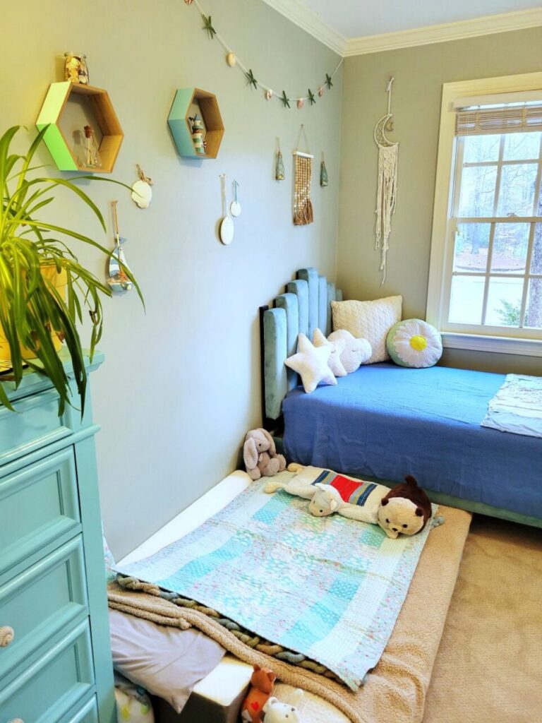 View of the children's bedroom (technically the kindergartener's room but where both boys now sleep at night). Straight ahead in far corner underneath a window (partially out of sight) is a twin bed and blue tufted headboard, covered in star-shaped throw pillows among other pillows and a blue blanket. To the immediate left (mostly out of sight) in corner by closet door is a blue-painted dresser with an airplane plant on top. On the floor in between the twin bed and dresser is a twin futon mattress tucked partially under the twin bed where the toddler sleeps, and is furnished with plush animals, several little pillows, and matching blanket. Several nautical decorations line the wall on the the left - hexagonal blue and teal shelves with mini lighthouse and glass bottle, jar of shells, hanging sailboat, sand dollars, etc. Above bed on window wall is a macrame crescent moon dream catcher