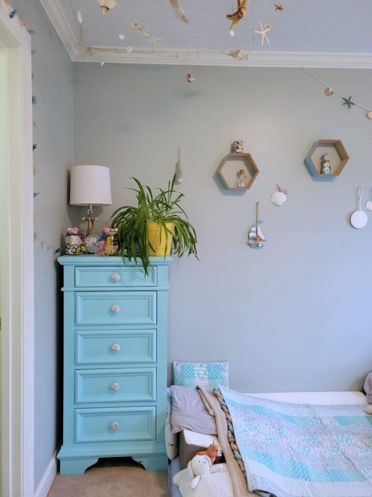 View of the children's bedroom (technically the kindergartener's room but where both boys now sleep at night) facing the wall with the blue-painted dresser. Atop it is an airplane plant, silver anchor-shaped lamp with white shade, clock, and other decorations. On the floor to the right is a twin futon mattress where the toddler sleeps (partially in sight) which is furnished with plush animals, several little pillows, and matching blanket. Several nautical decorations line the wall on the the left - hexagonal blue and teal shelves with mini lighthouse and glass bottle, jar of shells, hanging sailboat, sand dollars, etc. From the ceiling in the corner above the dresser is a fishing net with starfish and seashells 'caught' in it. To the left (mostly out of sight) is the open closet door. Strung above the closet doorframe is a strand of seashell lights.