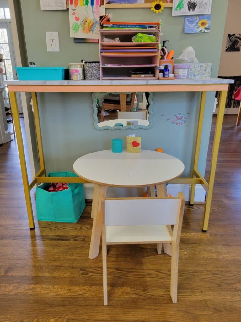 The console table is an efficient addition to the kitchen and dining room as it is positioned against a narrow wall separating the spaces and is tall enough to fit the toddler table underneath as seen in photo. Above is a bulletin board (partially seen) with colourful kids' artwork. On top is a five-tier wire rack for papers, a tissue box, a bin for miscellaneous items, and smaller bins for other designated items