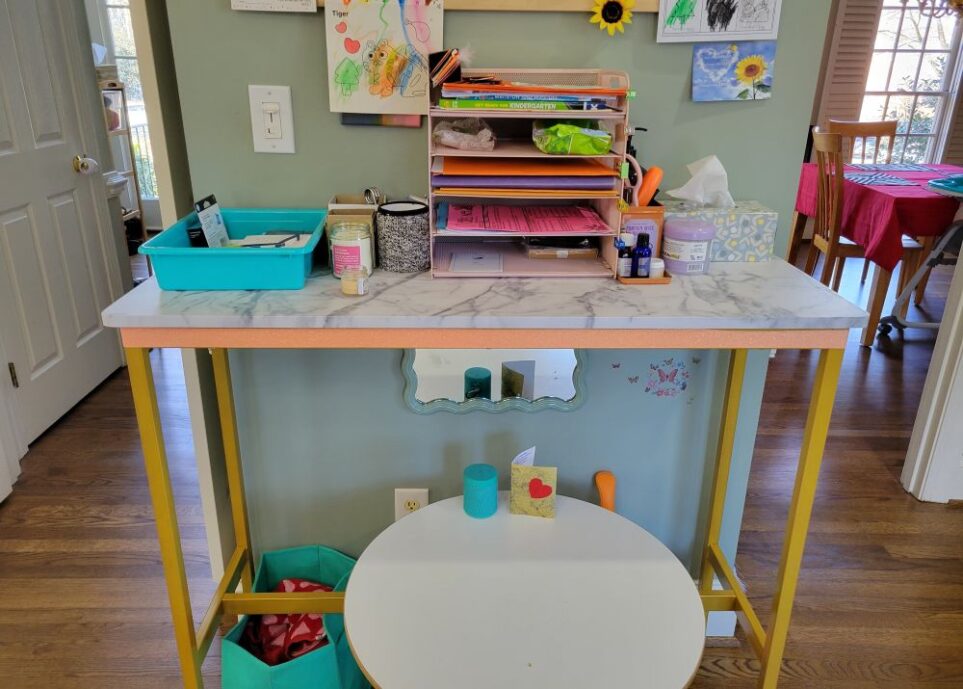 Console table in kitchen breakfast area, against a short span of wall between the dining room doorway (on right) and hallway leading into foyer (on left). Below it is a small white round toddler table. On the floor left of toddler table is a small teal cloth bin with soiled kitchen towels. Hanging on wall below console table, above the toddler table, is a sage-green-framed mirror. Along top of console table is a teal bin with items, small containers and a couple of candles, a pink five-tier paperwork metal tray with folders and other items organised in it, a tin filled with scissors, screw driver, etc. and a tissue box and other small containers. Above it hangs a bulletin board (mostly out of view) with children's artwork and schoolwork hanging on it.
