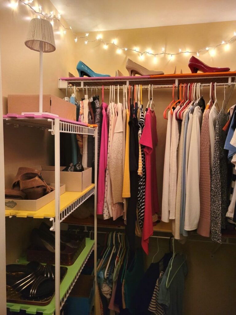 View of one corner of square-shaped walk-in primary closet: against back wall are shelves from which clothes hang and stop which three pairs of different-coloured pumps are displayed. Against left wall is a shelving unit lined in foam mats and filled with pairs of shoes on display in boxes and lids. On top is a small battery-powered white mini-lamp. Seen strung across ceiling at top is a lit strand of string lights