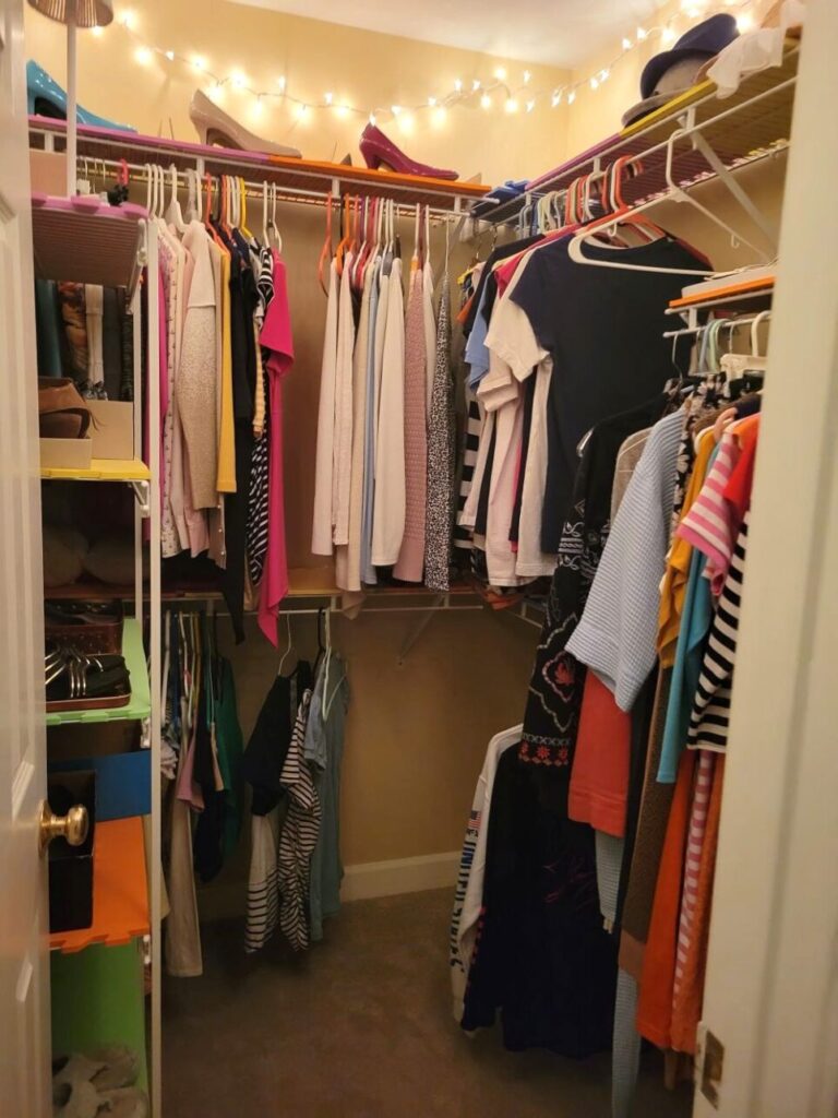 View of primary closet from open door: a square-shaped walk-in closet with shelves on right and back walls, from which clothes hang and stop which shoes and hats are displayed. Against left wall is a shelving unit lined in foam mats and filled with pairs of shoes on display in boxes and lids. On top (partially out of view) is a small battery-powered white mini-lamp