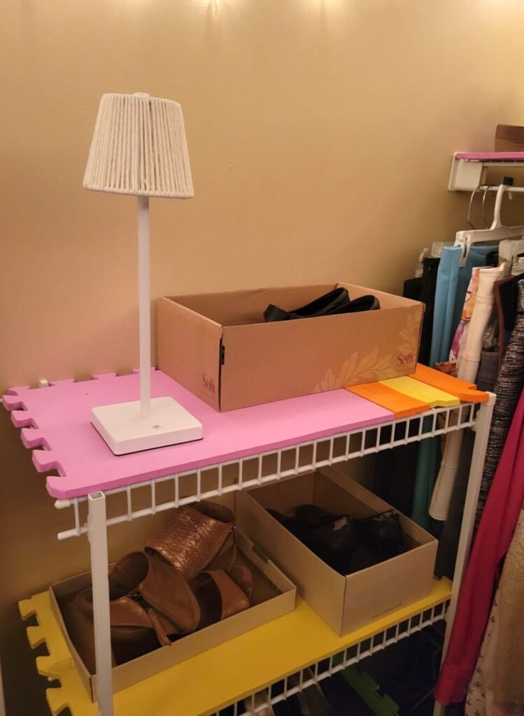 Top half of shelving unit seen in primary closet with a small battery-powered white mini-lamp next to a pair of heels in a shoe box on the top shelf. Below are another couple pairs of shoes on display in a shoebox and lid on the next shelf. The shelves are lined with foam mats