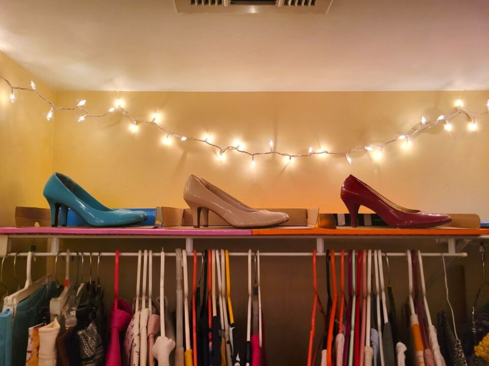 Close-up photo in primary closet of top shelf above clothes lined with foam mats and topped with three pairs of pumps in teal, nude and red, displayed atop partially-cut open shoe box lids. Above, lit string lights hang from the ceiling, casting a playful glow over the shoe display