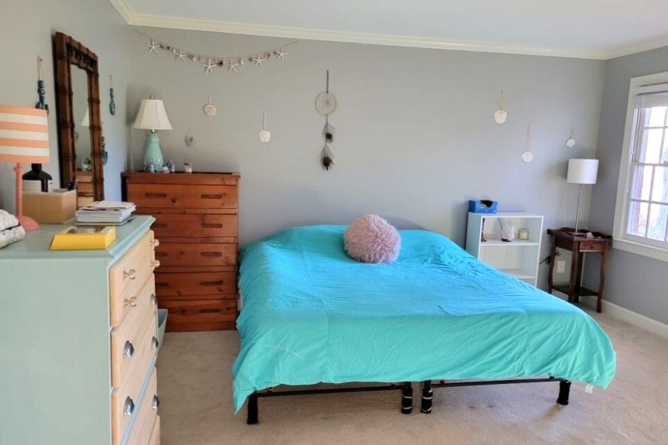 Primary bedroom with king bed in center against back wall, covered in bright blue comforter and shaggy pink throw pillow. Immediately to left in photo, against left wall, is a pale blue and wood dresser topped with small lamp and other organized items; a mirror and other decor hangs on wall beyond it, and a wood dresser stands in the corner next to bed topped with lamp and other decor. Small, simple decorations hang on back wall above bed and dresser and end tables. On right of bed is a small white shelved table with salt lamp, candle and tissue box, and to right of it in corner is small wood end table with lamp on top. To right of it against right wall is window (partially out of sight)