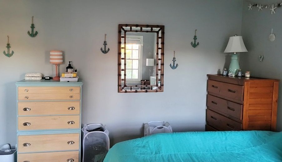 View of primary bedroom facing wall opposite windows from bed (partially in view). Toward left, closest to the door, is a pale blue and wood dresser topped with small lamp and neatly arranged personal items items. A wood-framed mirror hangs on wall to the right. Five small blue wooden anchors strung with twine hang in various places along the entire wall. In the right corner next to the bed is a wood dresser topped with a lamp and a few small nautical decorations. Along the wall above it hang a few small coastal decorations - seahorses, sand dollar, etc.