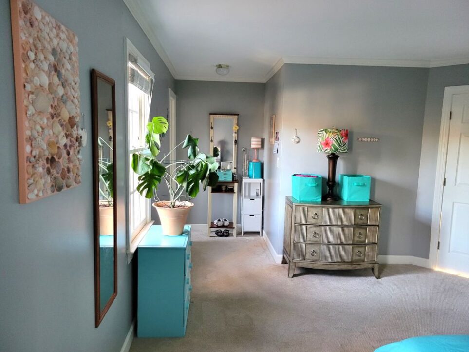 Primary bedroom viewed from back corner by bed (partially seen in photo): on right against opposite wall is brass-coloured dresser topped with a couple of blue cloth bins and a tall brown lamp in between with colourful tropical fern and pink flower lampshade. On wall are a couple of coastal decorations. Immediately left on wall hangs large seashell collage next to full-length mirror. Beyond it is window above blue-painted dresser and monstera plant on top. Further along wall in corner (barely in sight) is door to bathroom. To the right of bathroom door, in a small nook, are a standing vanity and small white end table with lamp on top, with jewelry box and various accessories on top