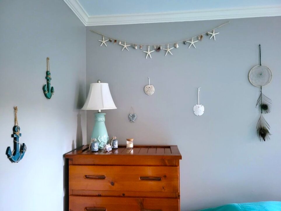Close-up photo of decor in corner of primary bedroom: top of wood dresser seen with blue seashell-carved porcelain-base lamp next to small coastal decorations and candle. On wall hang starfish/seashell garland, dreamcatcher above bed (barely seen to right side of image), a couple of sand dollars, and on the left wall a couple of blue wooden anchors strung with twine