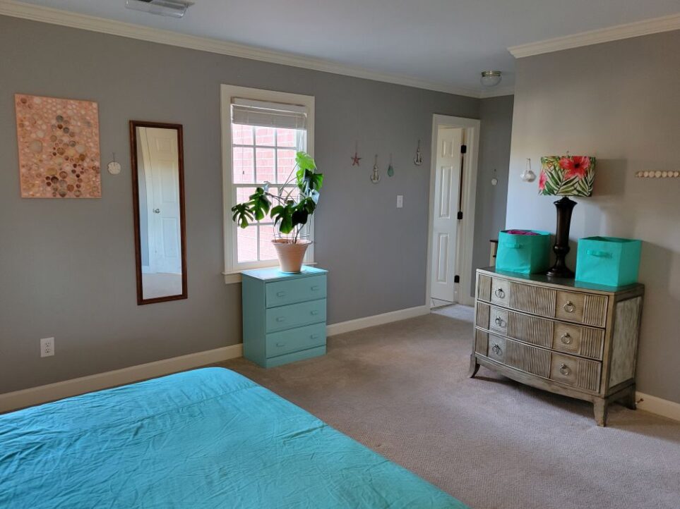 A corner of the primary bedroom viewed from bed (partially seen in photo): on right against wall is brass-coloured dresser topped with a couple of blue cloth bins and a tall brown lamp in between with colourful tropical fern and pink flower lampshade. On wall are a couple of coastal decorations. On left wall hangs large seashell collage next to full-length mirror and sand dollar. To right of it is window above blue-painted dresser with monstera plant on top. To right of that hang a few more nautical decorations on wall, and in corner is door to bathroom (standing vanity and additional lamp/end table are tucked into nook just out of line of sight)
