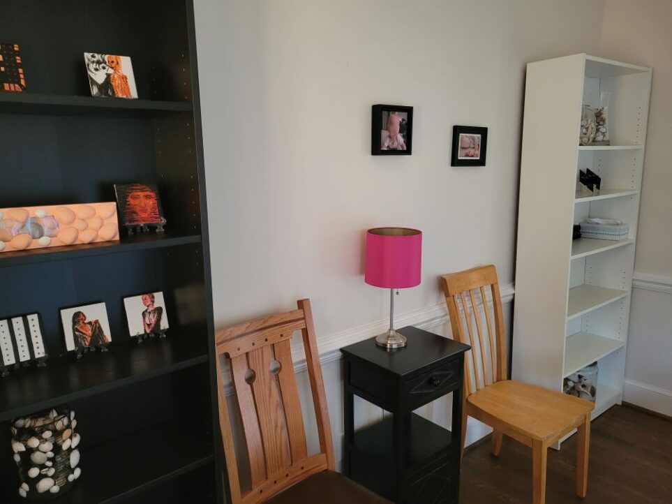 Dining room wall shown with black shelf on left (partially out of shot) and white shelf on right, with two chairs between and a small black end table between the chairs with pink lamp on top. Various painted tiles (with prominent colours of black, pink and orange) are on display on the shelves, and a couple of black-framed photographs hang on the wall above the chairs and table (in contrast to the white wall)