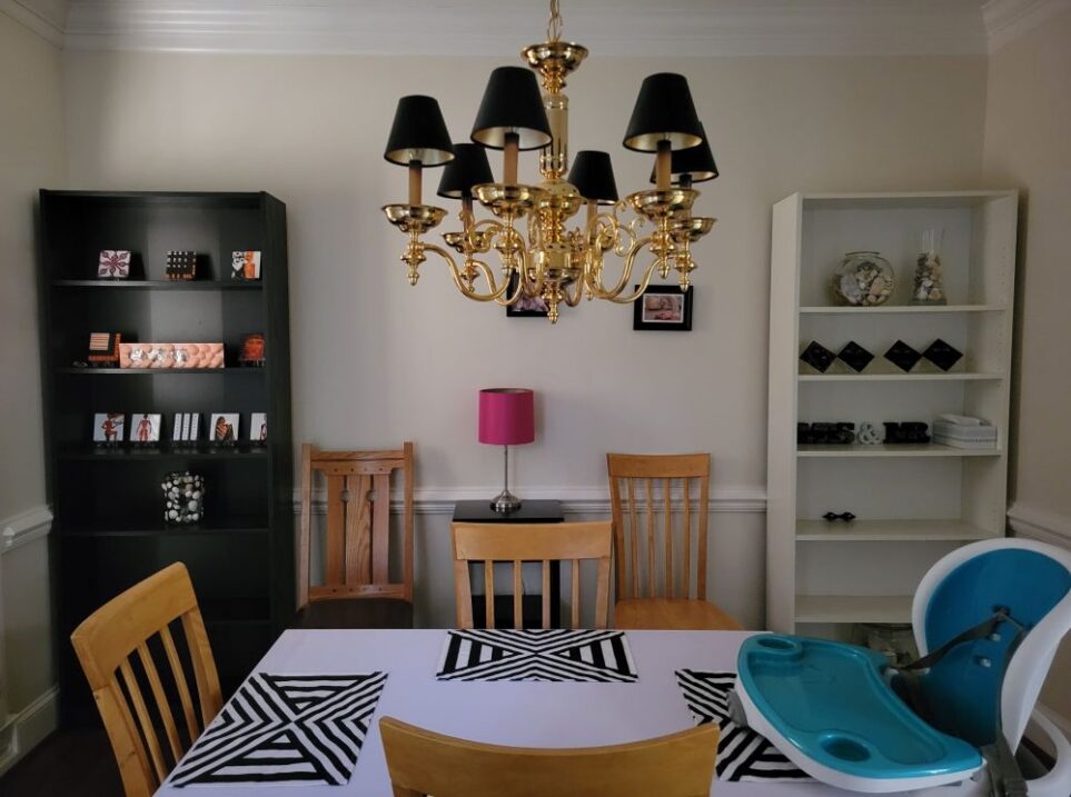 Dining room wall shown with black shelf on left and white shelf on right, with two chairs between and a small black end table between the chairs with pink lamp on top. Various painted tiles (with prominent colours of black, pink and orange) are on display on the shelves, and a couple of black-framed photographs hang on the wall above the chairs and table (in contrast to the white wall). In the center of the room is a table with three chairs and a high chair, below a candelabra-style gold-coloured chandelier with mini black lampshades. The table is covered in white peel-and-stick wallpaper on top and has four black and white geometric-patterned placemats