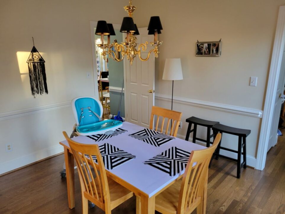 View of dining room facing door leading into kitchen. In the center of the room is a wood table with three chairs and a high chair, below a candelabra-style gold-coloured chandelier with mini black lampshades. The top of the table is covered in white peel-and-stick wallpaper and has four black and white geometric-patterned placemats. On the left wall hangs a black macrame evil eye design and on the right wall hangs a framed photograph collage. A black floor lamp with white shade stands next to open door and two black stools stand alongside wall to right of it, before the opening into the foyer