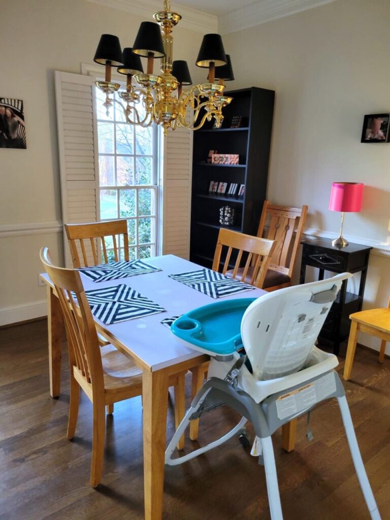 Dining room with table (top covered in white peel-and-stick wallpaper, as well as black and white geometric-design placemats) and highchair on one end, two additional chairs (partially out of view) lined against far wall with small black end table in between and pink lamp on top, gold-coloured candelabra-style chandelier with mini black lampshades, black bookcase in far corner with decor on shelves, and a couple of black-framed photos against the white walls on either side (window and open white plantation shutters to left of bookcase)