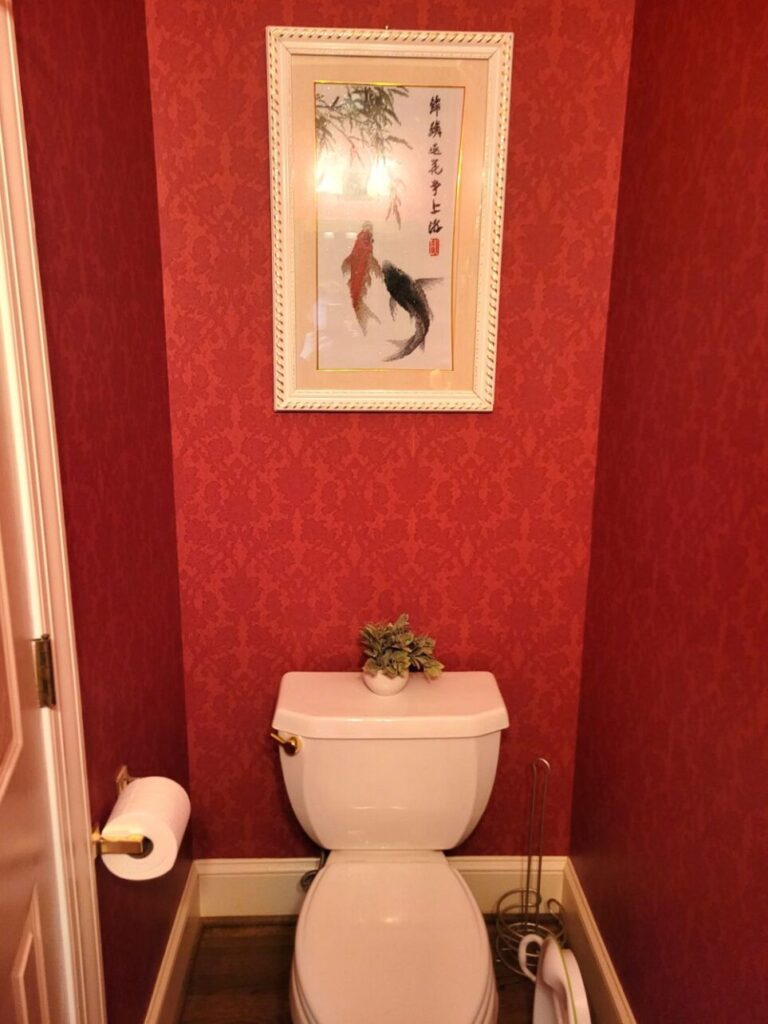 Power room viewed facing toilet: atop toilet is small artificial plant. A handmade framed knit picture of koi fish hangs on wall above. Walls are covered in red-patterned wallpaper