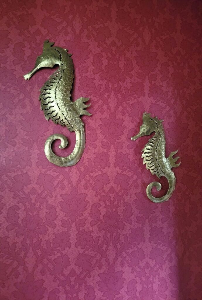 Two gold-painted metal seahorse sculptures hang on the wall in the powder room. Wall is covered in a dark red-print wallpaper