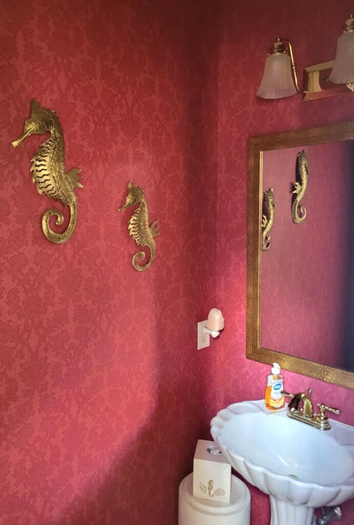 Power room viewed from door: a small scallop-shaped white porcelain sink stands to right side below a mirror in a gold-coloured dimpled frame. Below left of sink in the corner is a small upturned white plant stand repurposed as a table on which rests a seahorse-embossed tissue holder. On left wall hang two gold-painted metal seahorse sculptures. Walls are covered in a dark red-print wallpaper