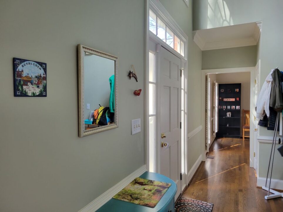 View of foyer from bottom of stairs, facing from the adjacent front living room. The blue table is seen in partial view below a mirror hung on the wall with some decor along either side, and just beyond is the front door. In far view ahead is dining room seen through doorway.