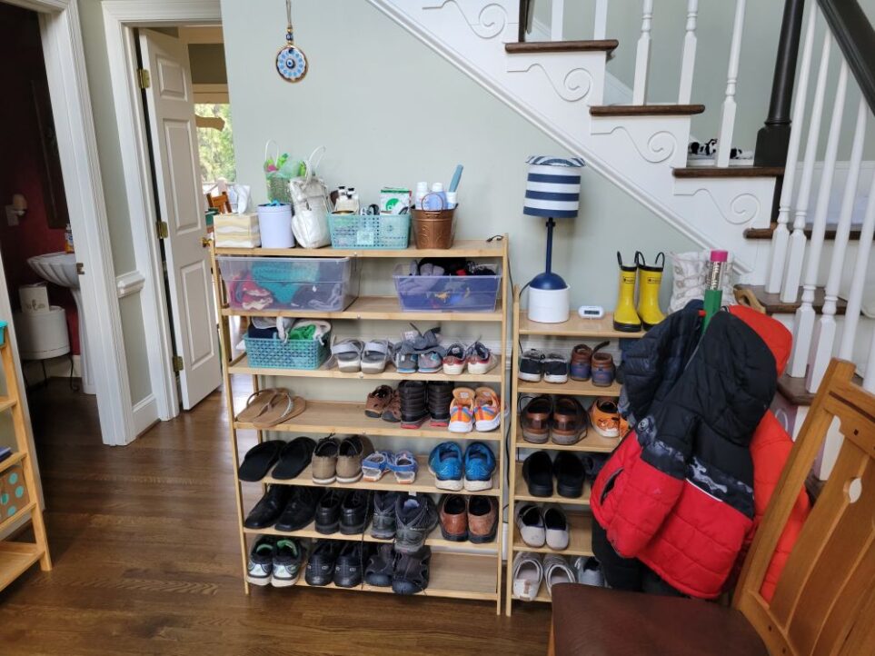 It's important to furnish a house with shoe receptacles like those shown in the foyer in this photo if you want to have a shoes-off house. To the left, a seven-tier shoe rack stands against the wall below the stairs next to a six-tier shoe rack. On the top shelves are various bins and everyday use items. A blue and white striped lamp stands atop the shoe rack on the right, alongside rainboots and a small clock. Both are filled with shoes on the lower shelves. In front of the shoe rack on the right is a child-sized coat rack hung with kids' coats, and in front of the coat rack is a chair (partially out of view) for taking shoes on/off