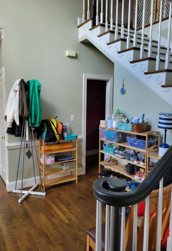 View of foyer and partial view of stairwell as seen from bottom of stairs. Seen across foyer are three shoe racks (one on opposite wall, next to coat rack) filled with shoes and with bags and other items organized on top