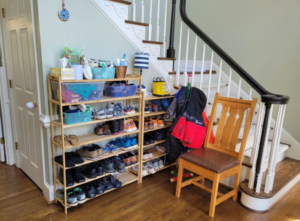 The stairwell 'nook' in the foyer: two shoe racks line the small wall before the stairs turn. A chair is located next to the base of the stairs, just inside the 'nook' and in between it and one of the shoe racks is a children's coat rack