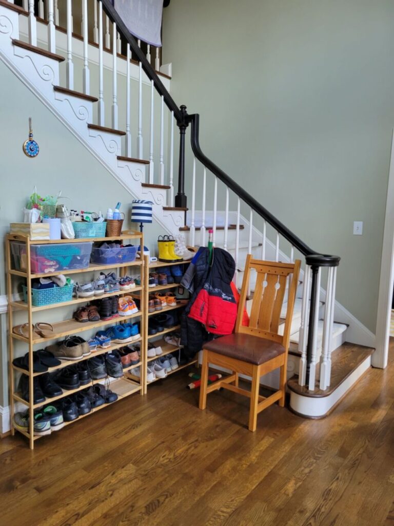 The bottom of the stairs open into the foyer, and below them is a 'nook' with a chair for removing shoes next to a children's coat rack and two tall shoe racks filled with shoes