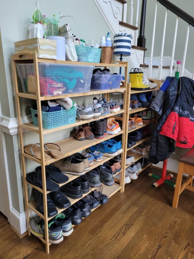In the foyer a seven-tier shoe rack stands against the stairwell wall next to a six-tier shoe rack. Both are filled with shoes as well as a few bins with winter accessories and everyday essentials. On one stands a small blue and white striped lamp. Adjacent to the smaller shoe rack in the corner of the stairwell nook is a child-sized coat rack with kids' coats, next to a chair for taking shoes on/off (most out of view)
