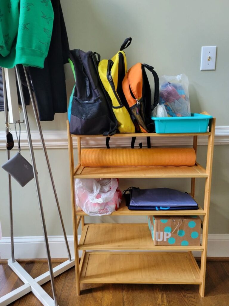 On the opposite foyer wall is a five-tier shoe rack with a couple of shoes (in a box/bag to give away), a yoga mat, a couple of empty bags, and on top are a few backpacks stocked and ready to go alongside a blue bin filled with extra toddler clothes, tissues, and other essentials ready-to-grab (furnish a house with convenient grab-and-go stations like these in mind). To the left of the shoe rack is a coat rack with some jackets hung at top out of sight.