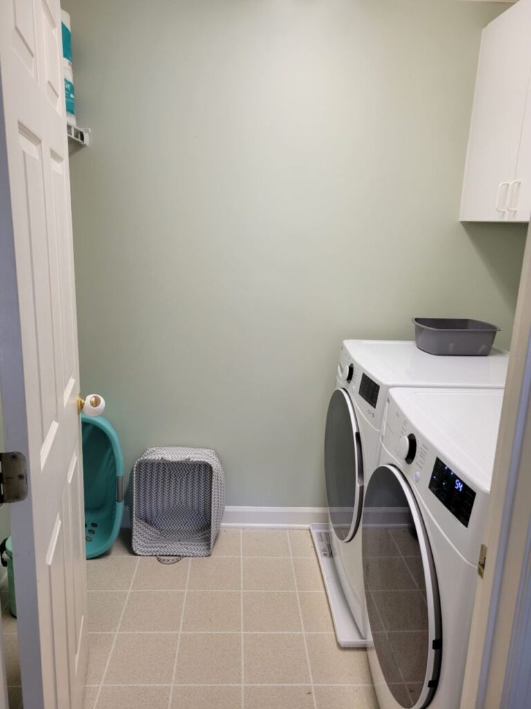 The laundry room as viewed from open door from upstairs hallway: to right are washer and dryer with white cabinets above (partially out of view). Up on the left wall, mostly out of sight on other side of open door, is a white rubber-coated wire shelf with an extra package of toilet paper on top. On the floor, standing upright on its side against the opposite wall, are two laundry baskets for clean loads.