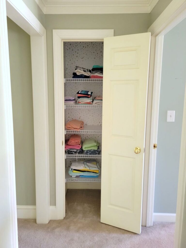 The linen closet by the children's bedroom viewed from the hallway. Its open door reveals several white rubber-coated wire shelves lined with a clear shelf-liner and filled with neatly folded towels and washcloths. The walls are covered in white peel-and-stick wallpaper with tiny gold stars and moons. To the right is the children's bedroom and to the left is the open hallway which continues down toward a bathroom, another closet, and the office