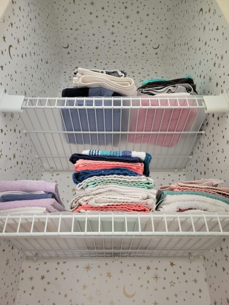 A view of the inside of the linen closet from below the top two shelves with folded washcloths. The walls and even the tiny square ceiling are covered in white peel-and-stick wallpaper with little gold moons and stars