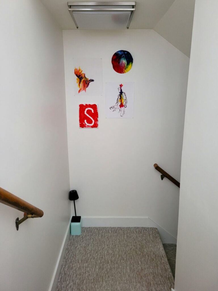 The basement stairwell landing, which turns to the right halfway down the stairs. A tiny black battery-powered mini-lamp rests atop a small upturned bin in the corner on the carpet, which can touch on at night when venturing down to the basement to avoid switching on the harsh fluorescent overhead light. On the wall hang four artworks: two homemade children's paintings and two professional prints from a street fair, all in vivid rainbow colours with prominent red and yellow