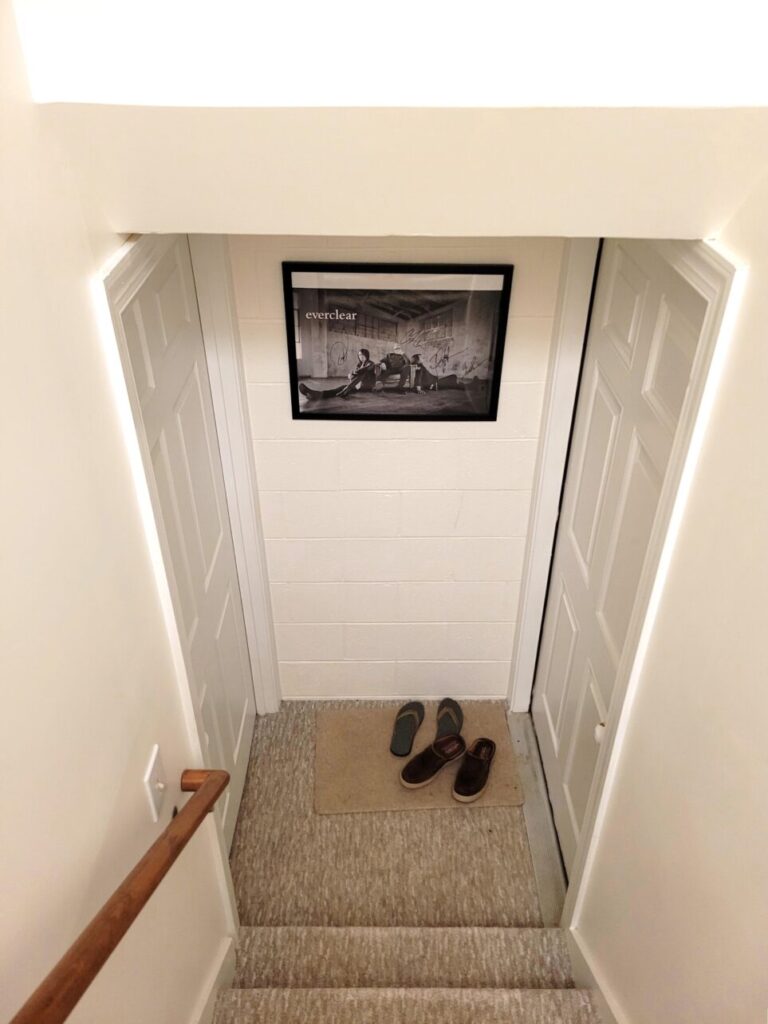 The bottom of the basement stairwell. To the right is the door leading into the garage. To the left is the door leading into the basement. A doormat is in the corner of the landing by the garage door with two pairs of shoes on it. On the white-painted cinderblock wall between the two doors is a black-framed photo of a black-and-white signed poster of Everclear