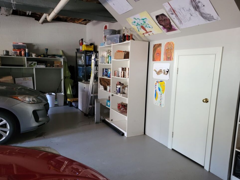 A portion of the back wall of the garage, which is covered with various paintings and drawings on the wall and slanted ceiling: a small white door leading into an under-the-stairs storage space toward the right, and shelving lining the wall down toward the left corner, filled with various organised items. A small chest freezer and another shelving unit fill the back corner, and more shelving continues along the next wall with various garage items. Two cars are seen parked in their spaces, mostly out of sight. Keeping a neatly stocked and tidy garage helps you furnish a house more easily because you can find things you are looking for without having to resort to storing things in the house itself due to a chaotic or messy garage.